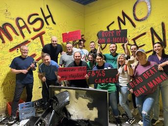 Group of adults posing in an indoor smash/rage room with bright yellow graffiti walls, holding sledgehammers and cheeky protest signs above a smashed TV and debris — lively team-building outing.