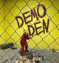 Person in red coveralls and yellow helmet smashing objects in a yellow-walled rage room behind a chain-link fence, scattered debris on the floor and large 'DEMO DEN' graffiti on the wall
