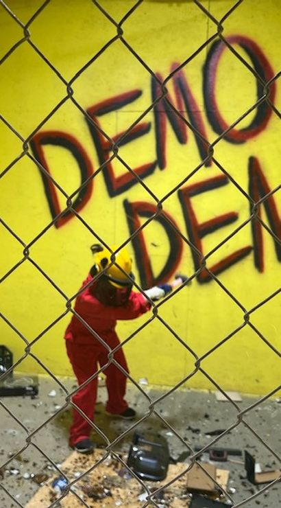 Person in red coveralls and yellow helmet smashing objects in a yellow-walled rage room behind a chain-link fence, scattered debris on the floor and large 'DEMO DEN' graffiti on the wall