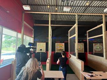 Indoor axe-throwing lanes with wooden targets behind safety cages in a red-walled entertainment space; a person tosses an axe while a camera operator records and a spectator captures it on a phone.