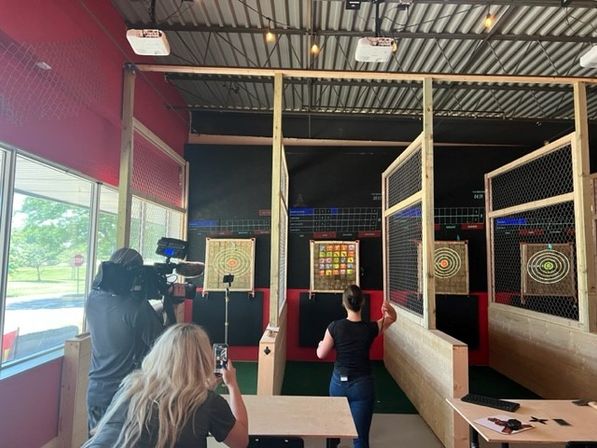 Indoor axe-throwing lanes with wooden targets behind safety cages in a red-walled entertainment space; a person tosses an axe while a camera operator records and a spectator captures it on a phone.