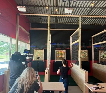 Indoor axe-throwing lanes with wooden targets behind safety cages in a red-walled entertainment space; a person tosses an axe while a camera operator records and a spectator captures it on a phone.