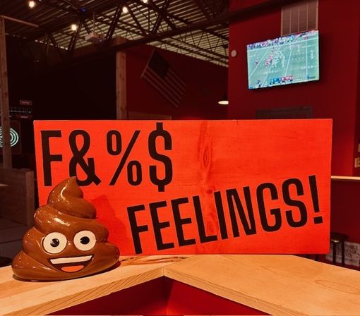 Orange wooden sign reading 'F&%$ FEELINGS!' with a smiling poop-emoji figurine on a wooden counter inside a sports-bar style interior with a TV showing football.