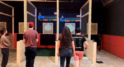 Three people from behind at an indoor axe-throwing range walking toward wooden target boards with illuminated bullseye rings, overhead electronic score screens, and fenced throwing lanes.