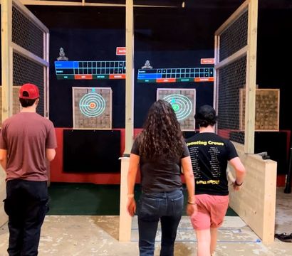 Three people from behind at an indoor axe-throwing range walking toward wooden target boards with illuminated bullseye rings, overhead electronic score screens, and fenced throwing lanes.
