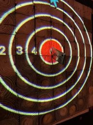 Glowing concentric target rings projected onto a wall of circular wood slices with a red bullseye and a small rectangular object lodged at the center, numbers 2-4 visible on outer rings.