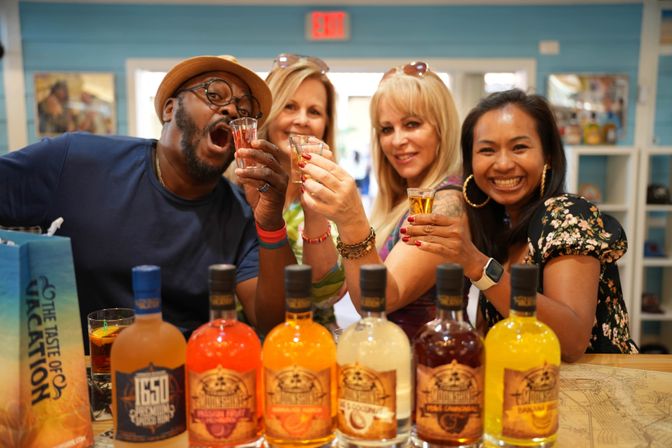 Four adults smiling and raising shot glasses behind a row of colorful spirits bottles in a bright tasting room, celebrating a lively bar-style tasting.