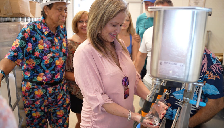 Woman filling a glass from a stainless-steel beverage filler on a small-batch craft brewery tasting tour, surrounded by people in colorful tropical shirts.