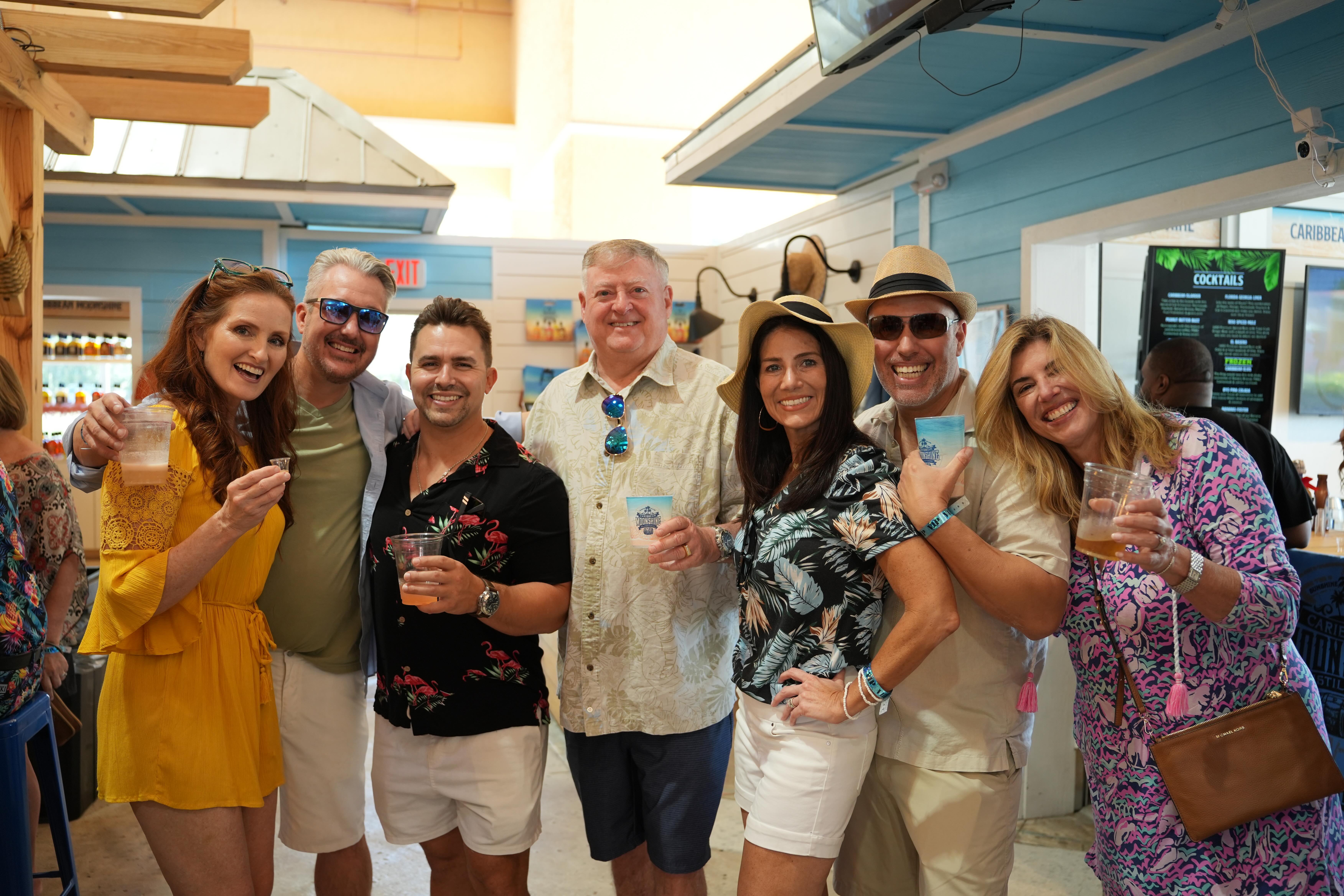 Seven smiling adults in summer outfits holding cocktails at a bright beach-themed bar with blue wood decor and sun hats.