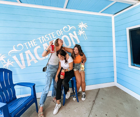 Three people laughing and holding bottled cocktails in front of a bright blue "The Taste of Vacation" mural with palm silhouettes and beach-style seating — summer coastal photo spot.