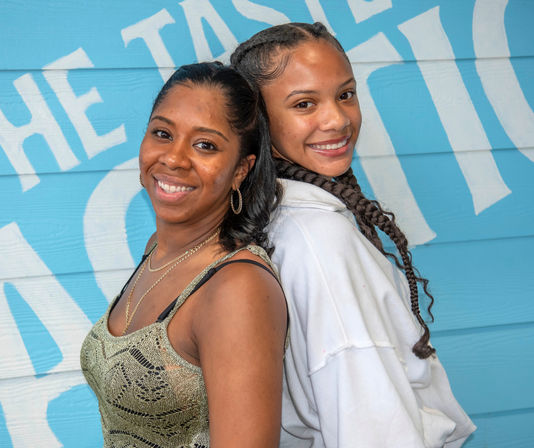 Two smiling women pose back-to-back against a bright blue mural — one with long braids in a white hoodie, the other in a patterned tank top and hoop earrings.