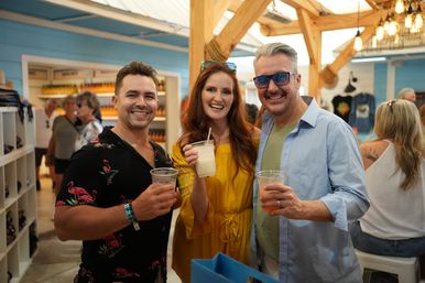 Three smiling adults raising cold drinks inside a bright beach-town souvenir shop with wooden beams and blue walls, vacation vibe