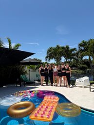 Seven women in black swimwear posing poolside on a sunny tropical backyard, palm trees and canal in the background, colorful inflatable floats bobbing in the blue pool.