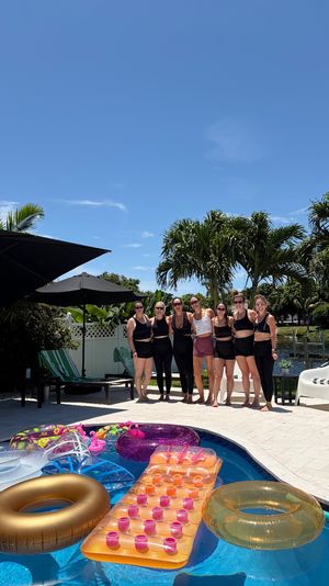 Seven women in black swimwear posing poolside on a sunny tropical backyard, palm trees and canal in the background, colorful inflatable floats bobbing in the blue pool.