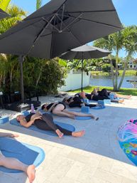 Relaxing outdoor yoga class on blue mats in a tropical waterfront backyard — participants lying under large black patio umbrellas beside palm trees and a canal.