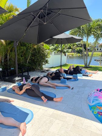 Relaxing outdoor yoga class on blue mats in a tropical waterfront backyard — participants lying under large black patio umbrellas beside palm trees and a canal.