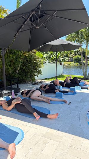 Relaxing outdoor yoga class on blue mats in a tropical waterfront backyard — participants lying under large black patio umbrellas beside palm trees and a canal.
