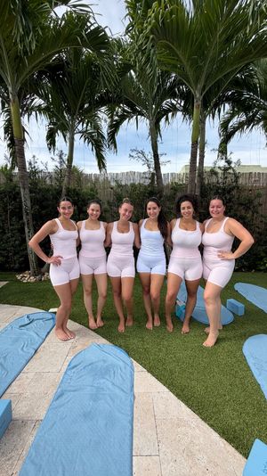 Six women in matching pale workout sets standing barefoot under palm trees in a tropical backyard, smiling in front of blue yoga mats and foam blocks arranged on a poolside patio.