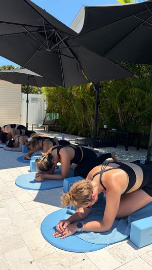 Group outdoor yoga class on a sunny stone patio — participants in child’s pose on round blue mats with foam blocks, shaded by large black umbrellas and tropical palms.