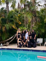 Group of nine friends in black activewear and sunglasses posing poolside in front of lush tropical palm trees and a hammock, beside a blue-tiled swimming pool with their reflections visible.