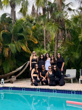 Group of nine friends in black activewear and sunglasses posing poolside in front of lush tropical palm trees and a hammock, beside a blue-tiled swimming pool with their reflections visible.