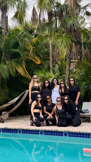 Group of nine friends in black activewear and sunglasses posing poolside in front of lush tropical palm trees and a hammock, beside a blue-tiled swimming pool with their reflections visible.