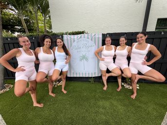Seven women in matching pale-pink and white activewear smiling and balancing in tree pose on artificial lawn in a tropical backyard, standing in front of a pastel striped banner with a palm graphic.