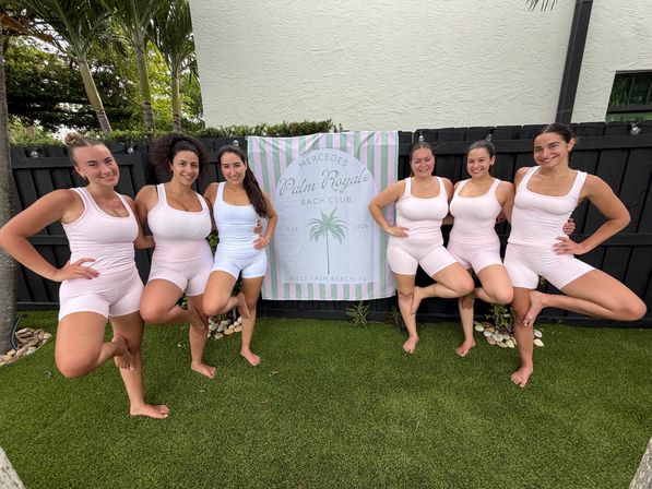 Seven women in matching pale-pink and white activewear smiling and balancing in tree pose on artificial lawn in a tropical backyard, standing in front of a pastel striped banner with a palm graphic.