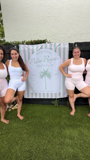 Seven women in matching pale-pink and white activewear smiling and balancing in tree pose on artificial lawn in a tropical backyard, standing in front of a pastel striped banner with a palm graphic.