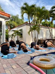 Five people in a poolside yoga class on blue mats and blocks beside a stucco house with red tile roof and palm trees, inflatable donut float and pink pool noodle in the pool.
