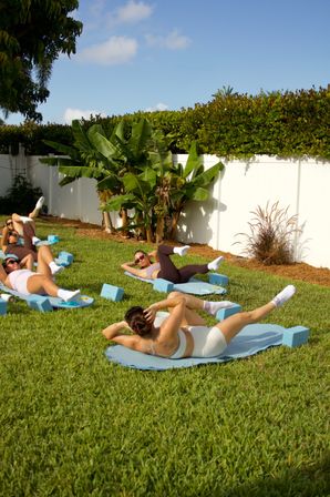 Sunny backyard fitness class with people doing mat Pilates and ab exercises on blue mats and foam blocks on a green lawn beside a white fence and banana plants