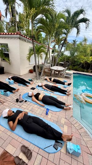 Group of six people resting on blue yoga mats beside a tropical backyard pool with palm trees, patio furniture and inflatable floats — outdoor relaxation session