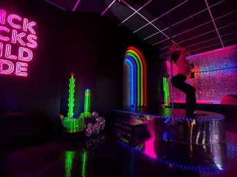 Vibrant neon-lit nightclub interior — silhouetted person on a pole on a mirrored disco-tile stage, rainbow LED arch, glowing neon cactus decorations, pink neon wall sign and glossy reflective floor.