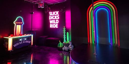 Neon-lit adult entertainment room with magenta glow, a suggestive novelty whack-a-game, glowing adult-themed slogan sign, disco balls and decorative cacti, rainbow LED arch and a small stage with a pole.
