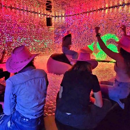Group of people in pink cowboy hats enjoying a neon-lit indoor ball pit with inflatable ride seats inside a shimmering sequin-walled art installation with colorful LED lights.
