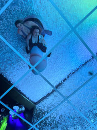 Two friends taking a mirror selfie while sitting on a pink inflatable seat inside a blue-lit indoor ball pit art installation with mirrored ceiling and sequin-covered walls.