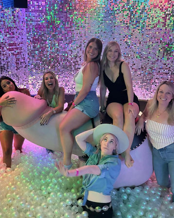 Group of friends laughing and posing in an indoor interactive art installation with an iridescent sequin wall, LED color lighting, a large white curved sculpture, and a glowing pearl-white ball pit — playful party photo in casual summer outfits.
