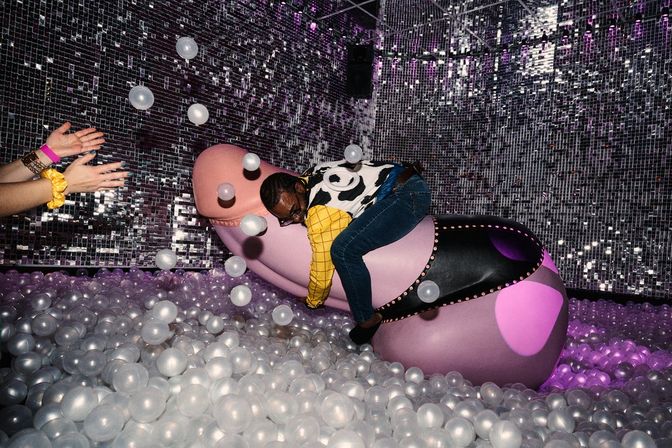 Person climbing a pink sculptural seat in an immersive indoor art installation with mirrored disco walls and a sea of translucent ball-pit balls