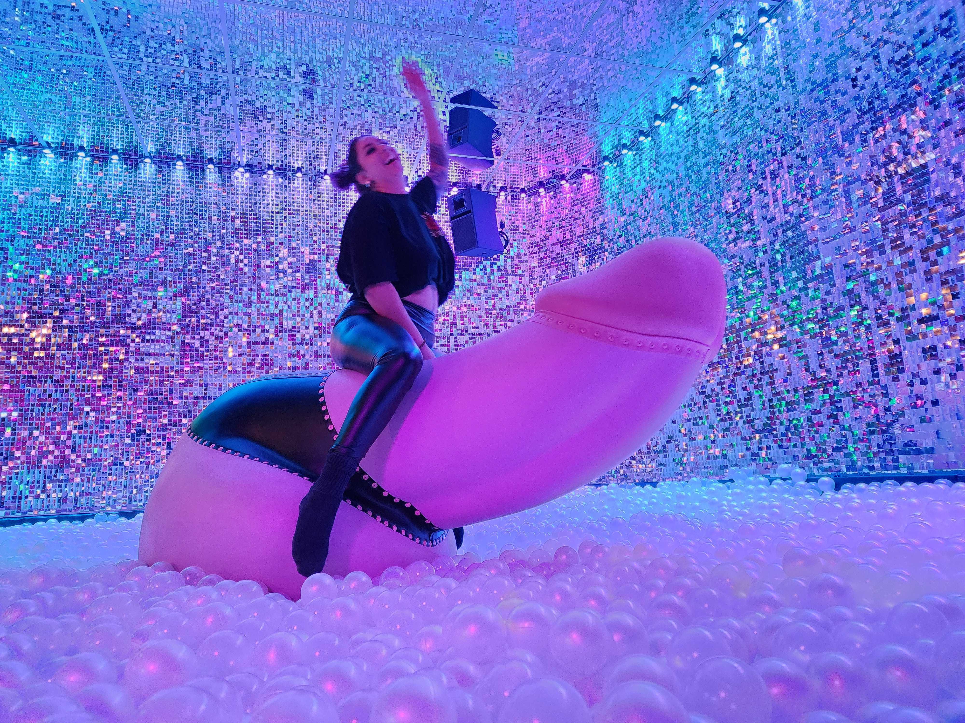 Person in black outfit riding a giant pink phallic sculpture inside a neon-lit interactive art installation ball pit with mirrored sequin walls and multicolored LED lights.