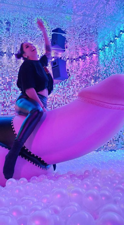 Person in black outfit riding a giant pink phallic sculpture inside a neon-lit interactive art installation ball pit with mirrored sequin walls and multicolored LED lights.