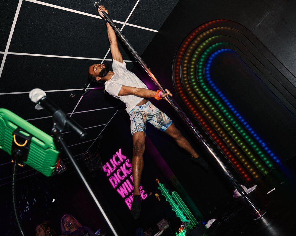 Male pole dancer mid-spin on a chrome pole in a neon-lit nightclub stage with rainbow LED arch and pink neon sign, wearing a white tee and patterned shorts, microphone stand in foreground.