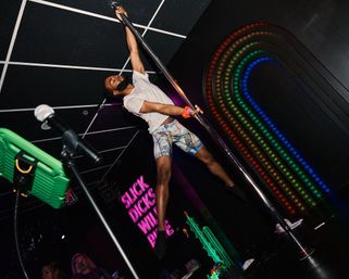 Male pole dancer mid-spin on a chrome pole in a neon-lit nightclub stage with rainbow LED arch and pink neon sign, wearing a white tee and patterned shorts, microphone stand in foreground.