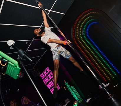 Male pole dancer mid-spin on a chrome pole in a neon-lit nightclub stage with rainbow LED arch and pink neon sign, wearing a white tee and patterned shorts, microphone stand in foreground.