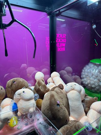 Arcade claw machine filled with novelty plush penis toys in assorted skin tones, clear prize capsules, and a neon purple backdrop