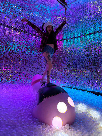 Person in a cowboy hat balancing on an oversized shoe sculpture in an immersive mirrored LED room over a white ball pit, vibrant purple and blue neon lights and a visible tattooed leg.