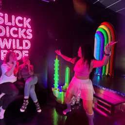 Neon-lit lounge scene with three friends: one dancing in a pink crop top and pleated skirt in front of a rainbow LED arch, two seated sipping drinks beside a bright pink neon wall sign and glowing green sculptural lights.