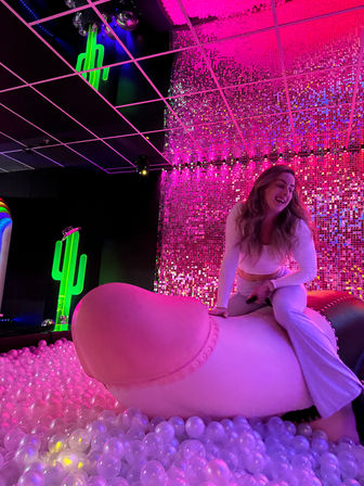 Smiling woman in white outfit perched on a giant pink novelty sculpture in a neon-lit indoor art exhibit with a pink sequin wall, clear ball pit and green neon cactus sign