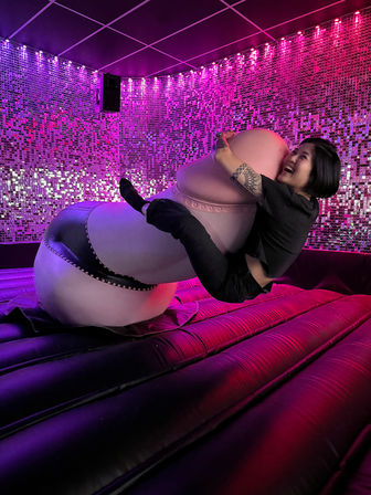 Person laughing while hugging an oversized pink novelty prop on a black inflatable mattress in a neon-lit lounge with sparkling sequin wall and purple-pink LED lights.