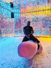 Adult in a black shirt sitting on an oversized pink sculpture inside a colorful immersive art installation with mirrored sequin walls, rainbow LED lighting and a white ball-pit floor.