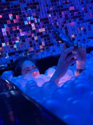Person peeking from a neon blue and purple illuminated ball pit, taking a smartphone selfie in front of a mirrored mosaic wall at an indoor interactive art installation.
